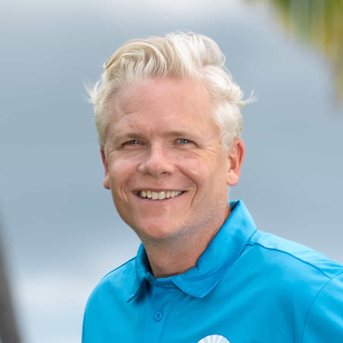 A man in a blue shirt smiling in front of palm trees.